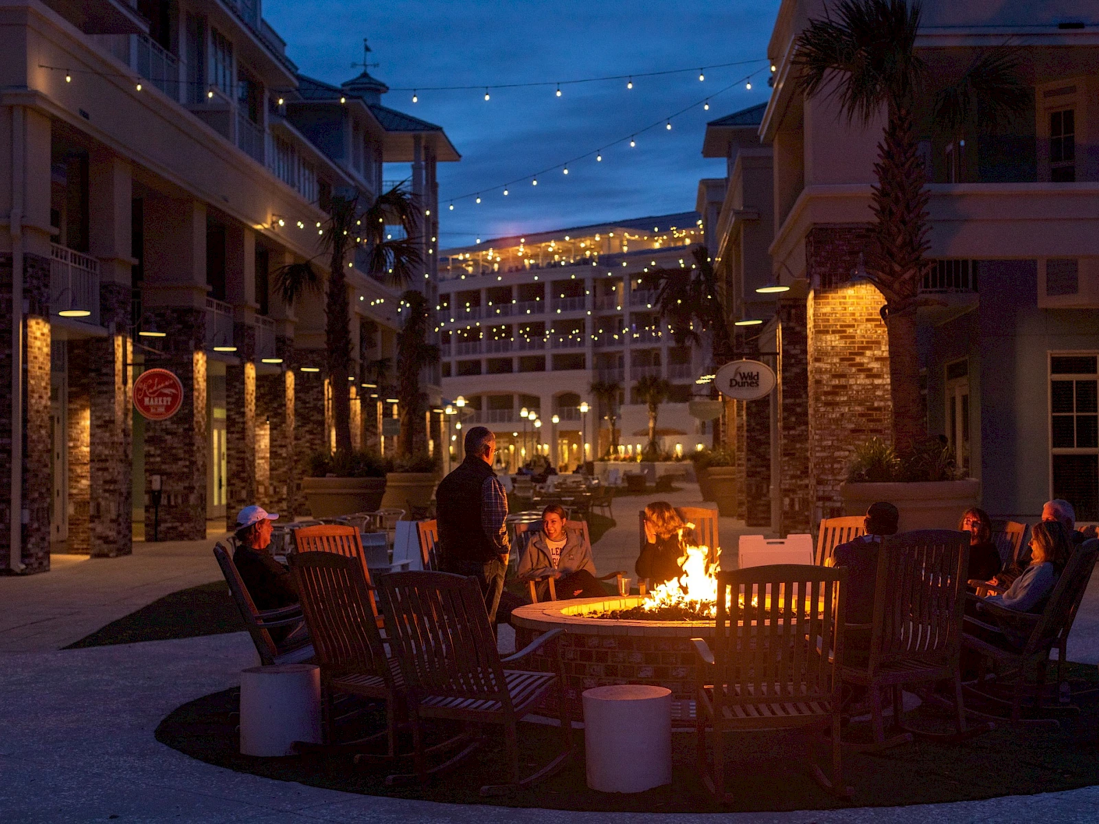 People gather around a glowing fire pit in an outdoor plaza at dusk, string lights overhead, cozy seating, and warm conversation continuing late into the night.