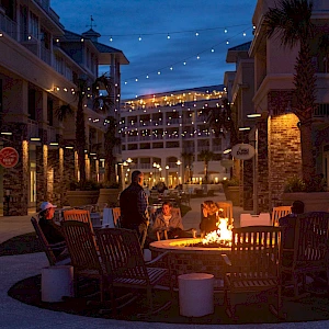 People gather around a glowing fire pit in an outdoor plaza at dusk, string lights overhead, cozy seating, and warm conversation continuing late into the night.