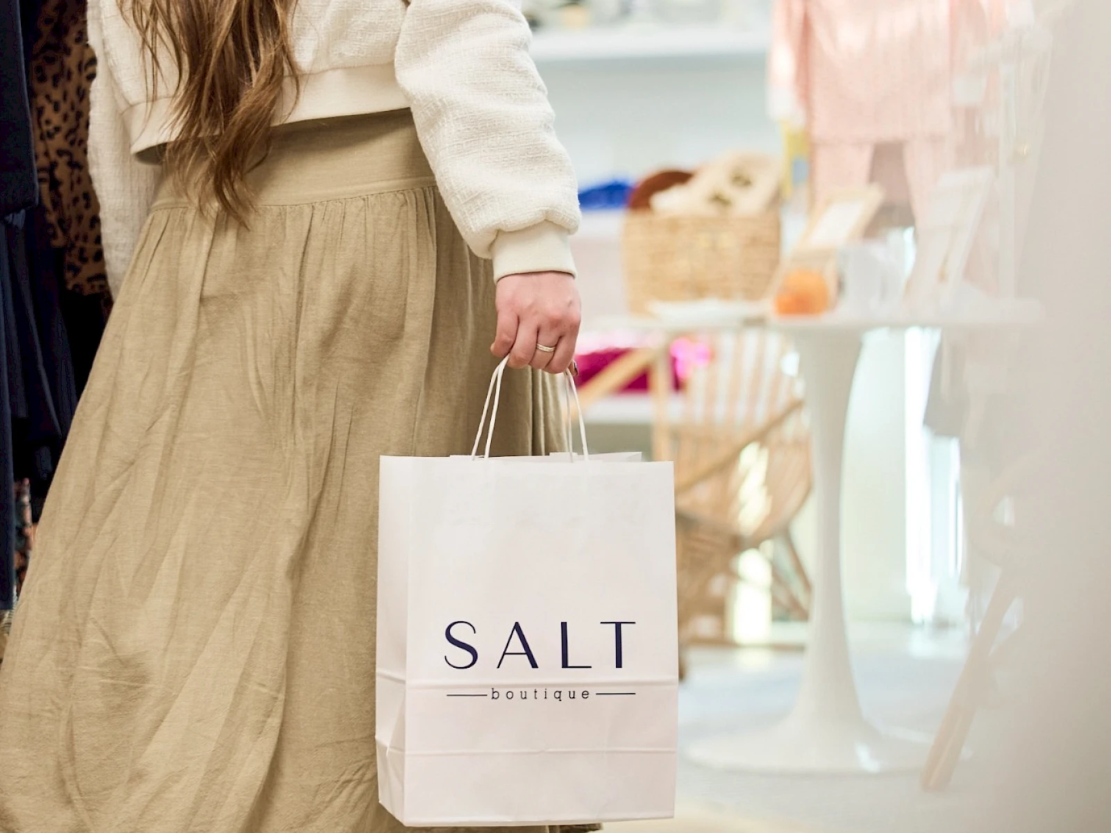 A woman in a beige skirt and white jacket walks through a bright store, carrying a white bag labeled “SALT” as shelves glow with pastel displays.