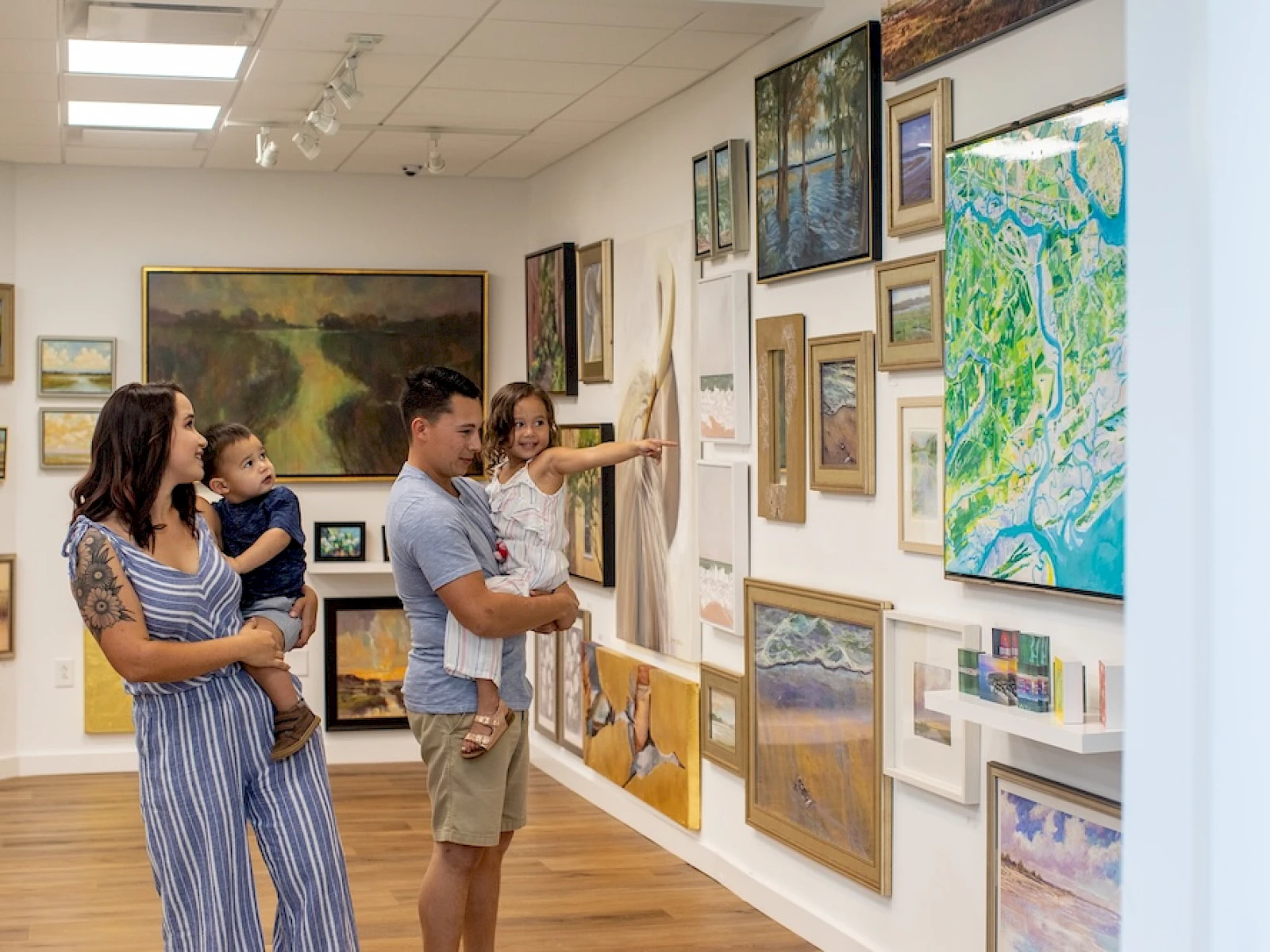 A family of four browses an art gallery, a man points to a wall of paintings and maps while a woman holds a child and smiles, exploring together.