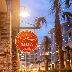 A red circular sign for a market hangs by a brick storefront at dusk, palm trees, string lights, and a sidewalk cafe vibe.