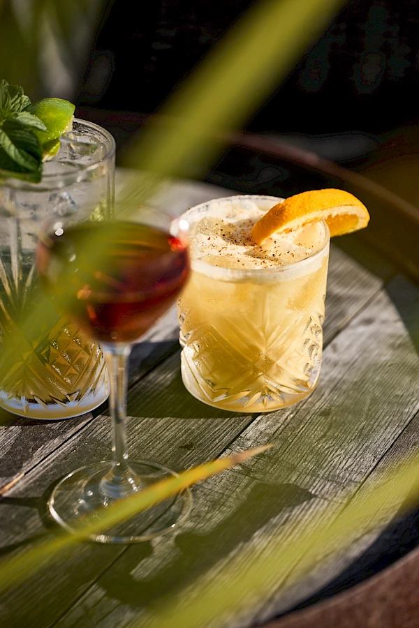 Three cocktails on a wooden table: a light-colored drink with an orange slice, a red wine glass, and a minty-looking beverage in the background, all under soft lighting.