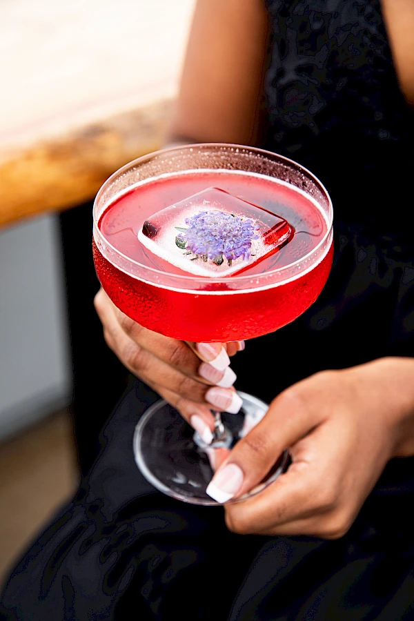 A stylish pink cocktail in a coupe glass, garnished with a lavender flower, held by a person in a black dress.