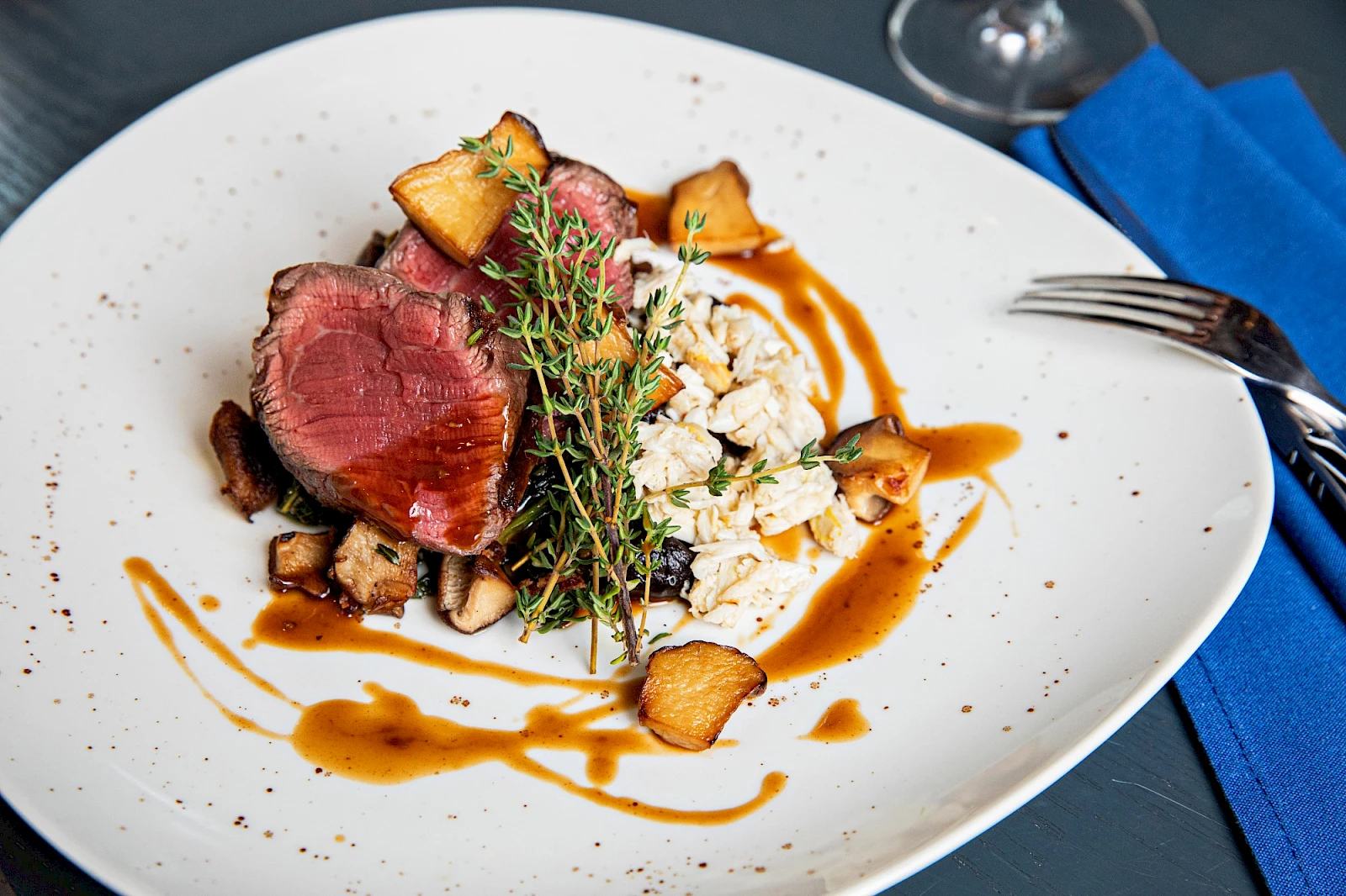 A plated gourmet dish: sliced seared steak, crispy croutons, grains, a sprig of thyme, and a caramel sauce drizzle on a white plate. Ends.