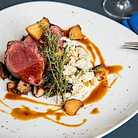 A plated gourmet dish: sliced seared steak, crispy croutons, grains, a sprig of thyme, and a caramel sauce drizzle on a white plate. Ends.