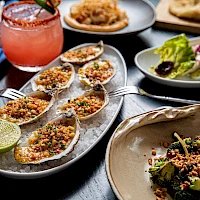 A spread of appetizers: toasted oysters on the half shell topped with crunchy herbs, lime wedges, a bright cocktail, and a fresh salad.