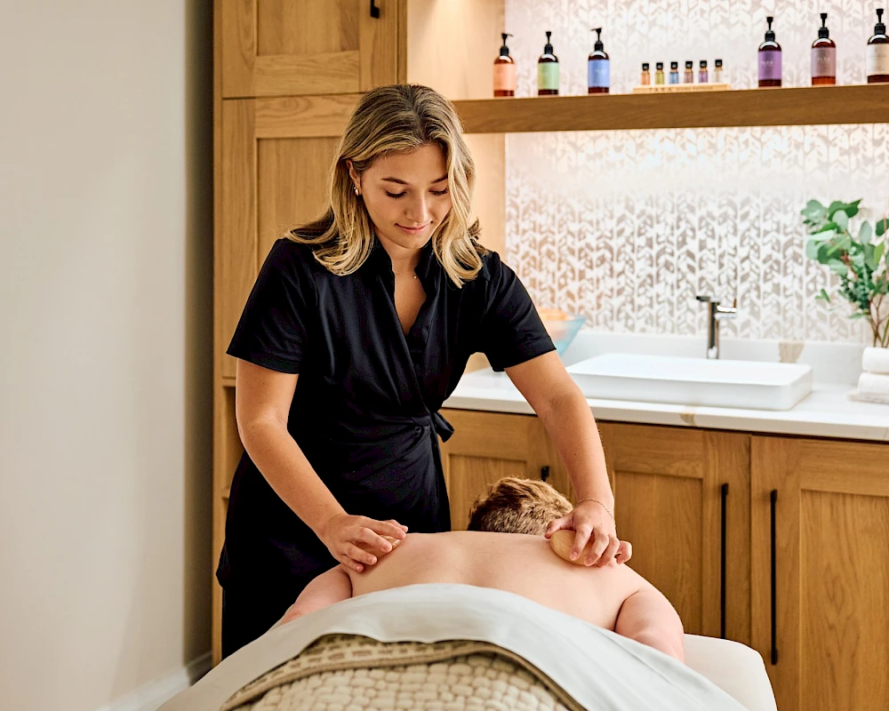 A spa scene with a therapist giving a facial or head massage to a client lying face down on a treatment bed, in a calm, wood-accented room.