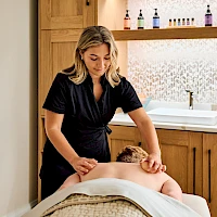 A spa scene with a therapist giving a facial or head massage to a client lying face down on a treatment bed, in a calm, wood-accented room.