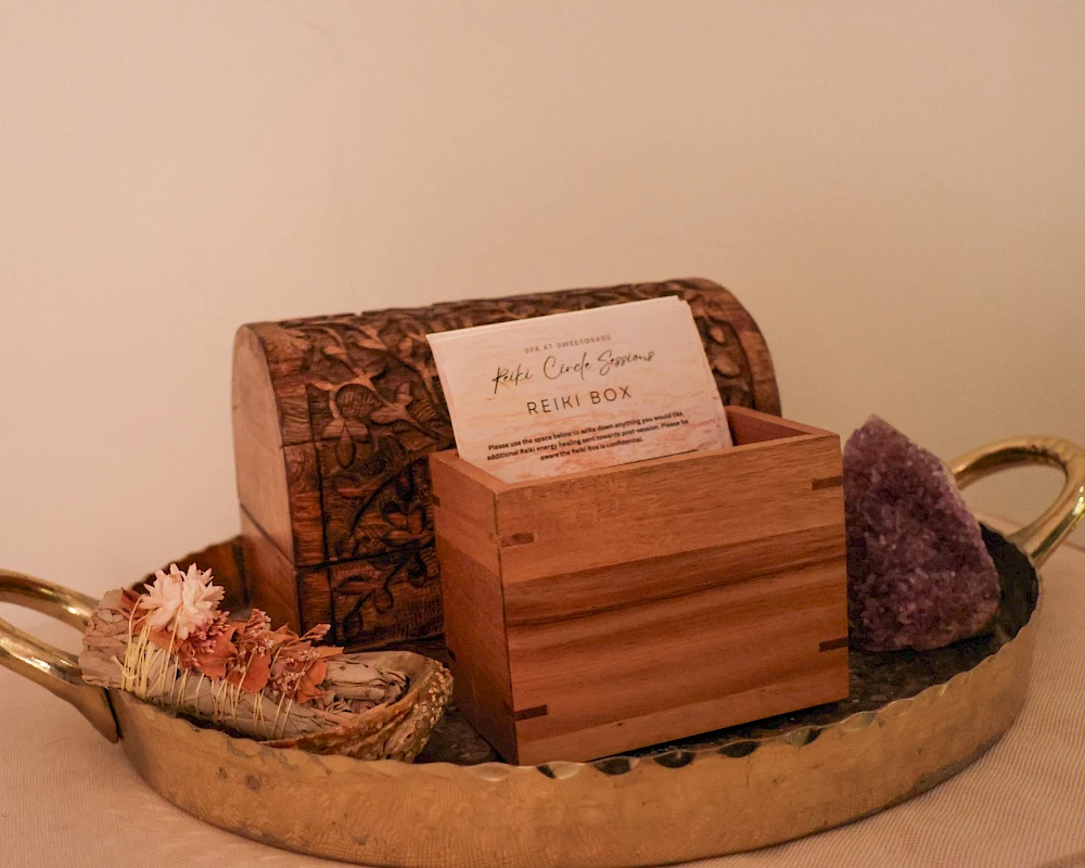 A round metal tray with two gold handles holds a wooden box, a carved wooden case, a bar of soap, and a purple crystal cluster on a beige background.