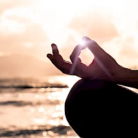 A pregnant person at the beach makes a hand gesture framing the sun, creating a serene sunset moment with soft waves in the background.