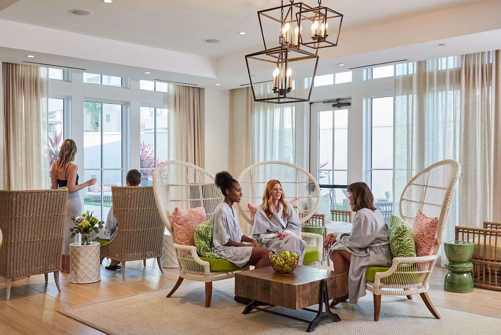 A bright lounge with wicker chairs and a coffee table, four women chatting in robes, sunlight streaming through large windows, cozy plants and modern hanging lights.