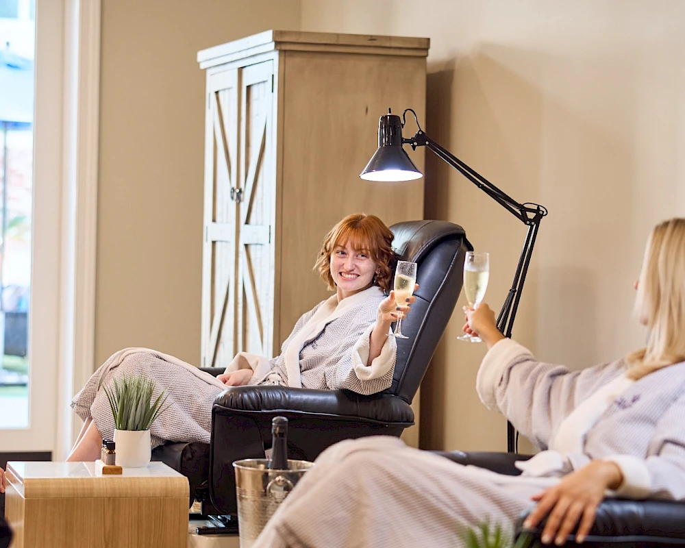 People relaxing in a cozy spa or salon: a woman sits in a comfy chair under a lamp, another person feet up, and a foot soak station with plants nearby.