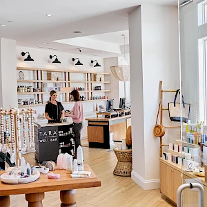 A bright boutique with shelves, display tables, and two customers browsing near a checkout counter, natural light streaming through large windows.