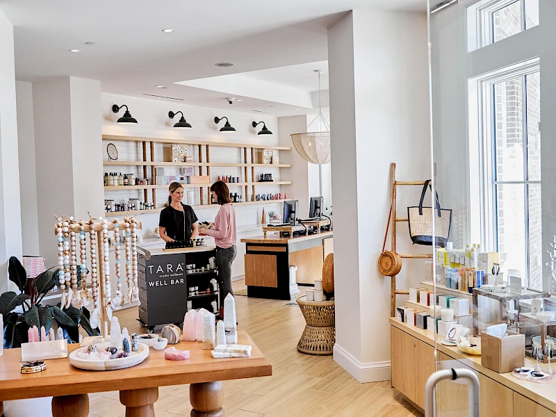 A bright boutique with shelves, display tables, and two customers browsing near a checkout counter, natural light streaming through large windows.