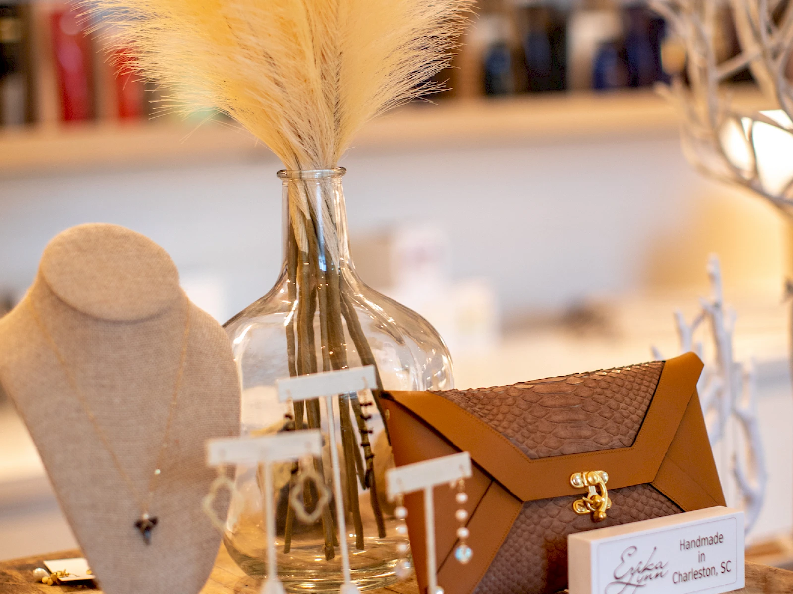 A cozy craft setup on a wooden table with dried bouquet, white display stands, and a small brown box, nicely arranged against a blurred shop backdrop.
