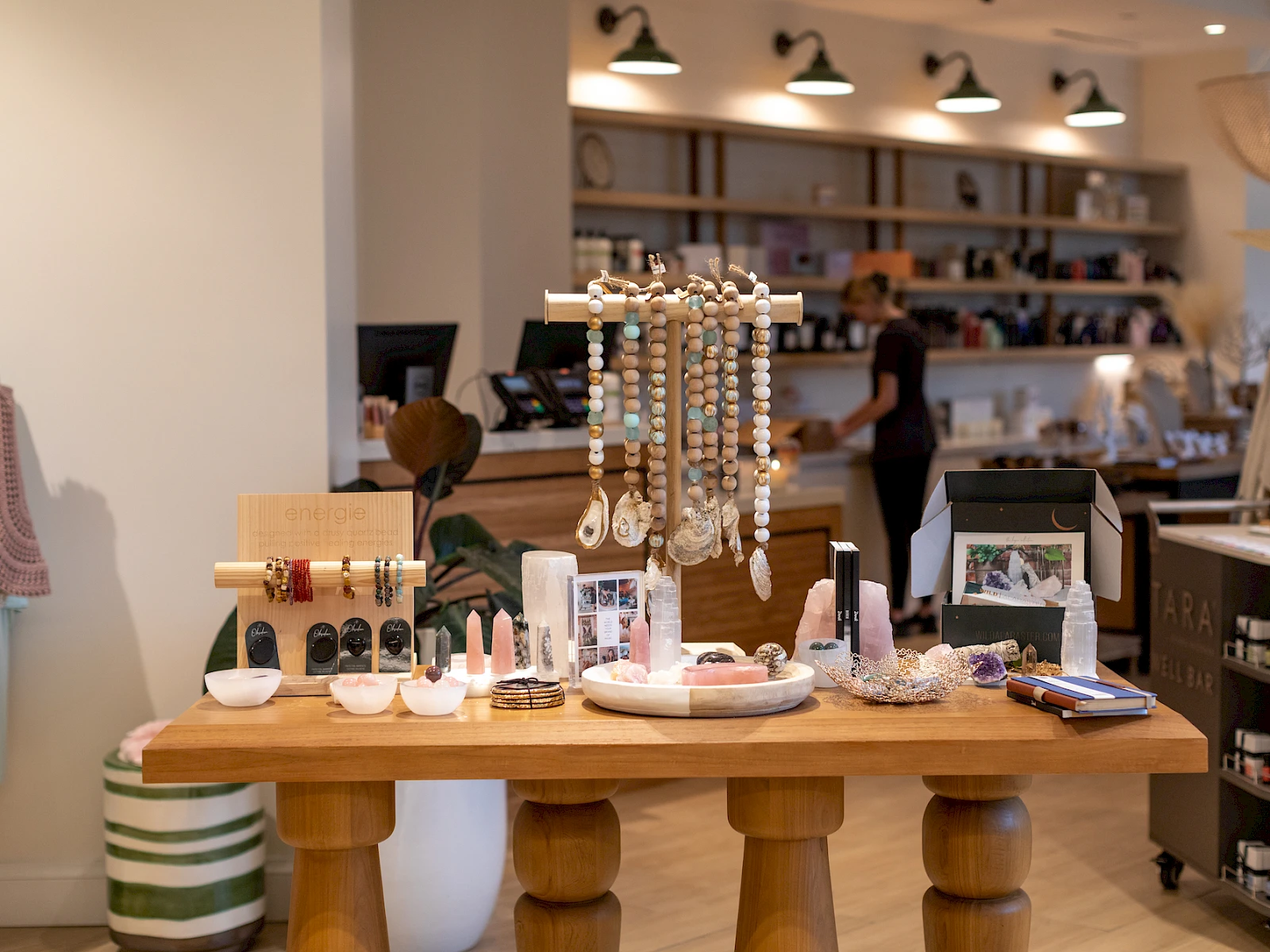 A cozy boutique shop with a wooden display table featuring jewelry, accessories, and decor; shelves line the back, shoppers browse softly.