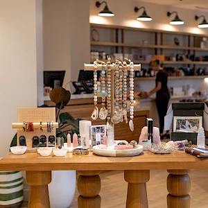 A cozy boutique shop with a wooden display table featuring jewelry, accessories, and decor; shelves line the back, shoppers browse softly.