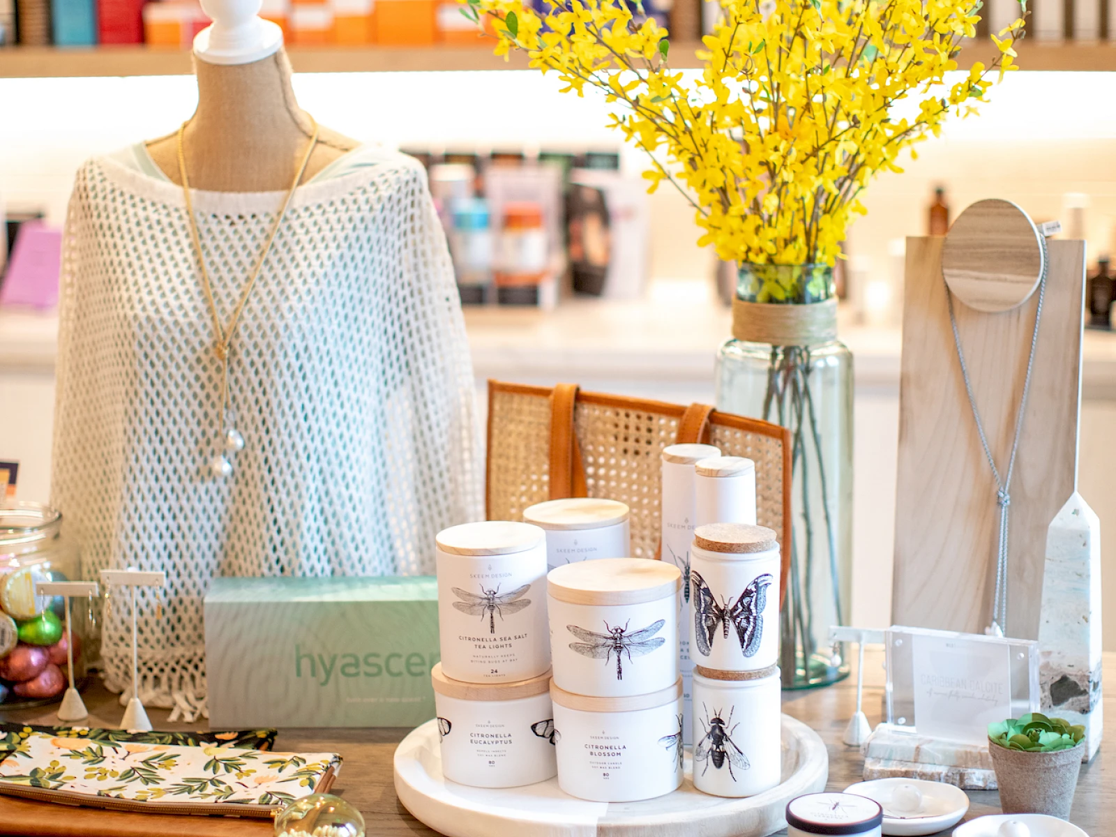 A chic boutique display featuring skincare jars, a white knit sweater on a mannequin, yellow flowers, and shelves of colorful products in the background.