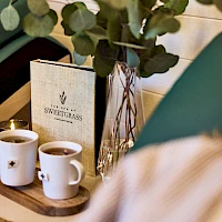 Two white mugs sit on a round tray, steam rising from coffee, next to a menu and a vase of eucalyptus, in a cozy cafe setup.