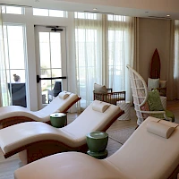 A bright spa lounge with curved white loungers, green bedside pots, sheer curtains, and a relaxed seating area by glass doors.