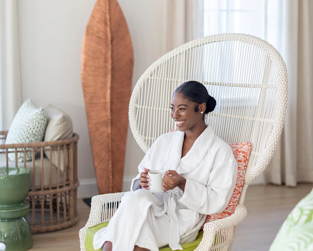 A woman in a white robe sits in a white wicker chair indoors, smiling while holding a mug; tropical decor and large window backdrop.