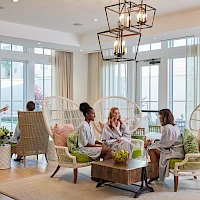 A bright, airy lounge with wicker chairs, four people chatting in robes, a coffee table with plants, and large windows with sheer curtains.