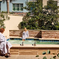 Two people sit by a shallow pool in robes, chatting beside a brick wall and lush green trees near a sunny courtyard.