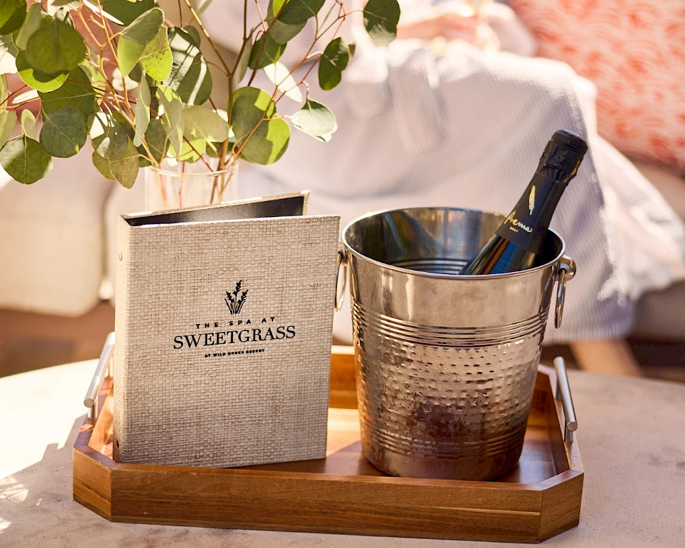 A small wooden tray with a plant and a metal ice bucket sits on a table; a cozy outdoor scene with soft cushions in the background.
