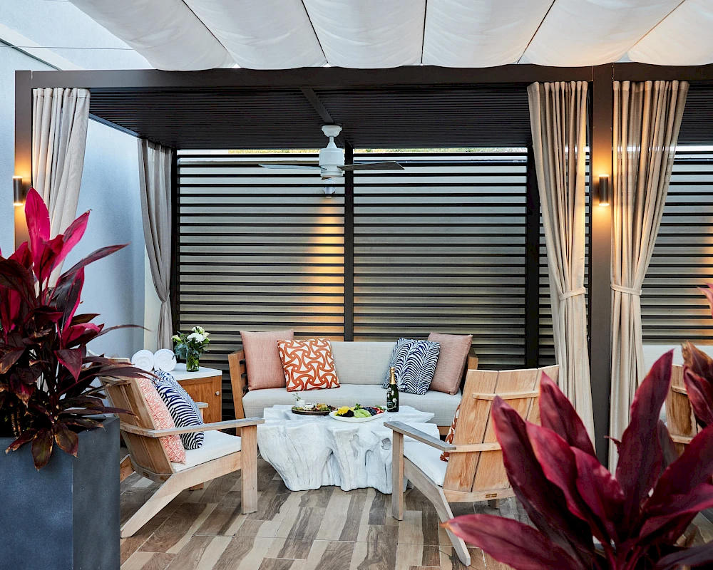 A stylish outdoor lounge with wicker chairs, a white coffee table, cozy cushions, potted plants, and draped curtains under a shaded pergola.