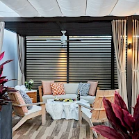 A stylish outdoor lounge with wicker chairs, a white coffee table, cozy cushions, potted plants, and draped curtains under a shaded pergola.