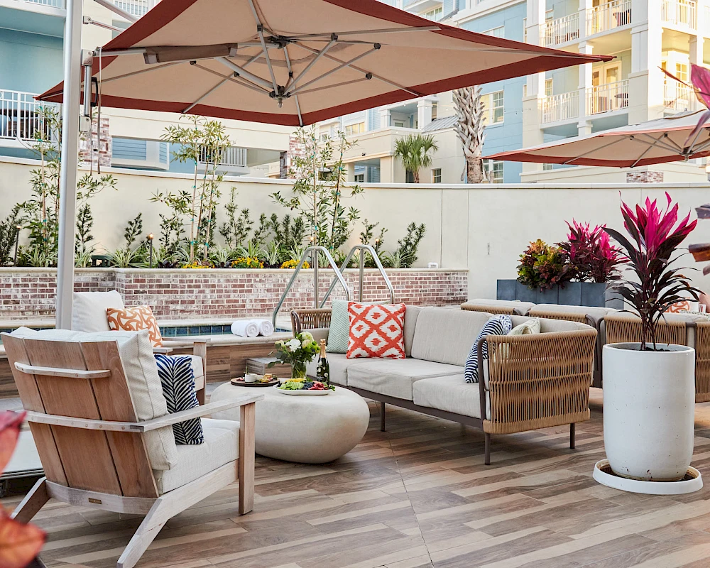 A sunny outdoor lounge with beige wicker sofas, colorful cushions, round coffee tables, potted plants, and a large umbrella shading the seating area.