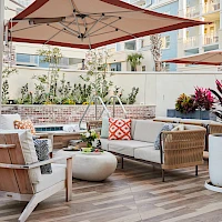 A sunny outdoor lounge with beige wicker sofas, colorful cushions, round coffee tables, potted plants, and a large umbrella shading the seating area.