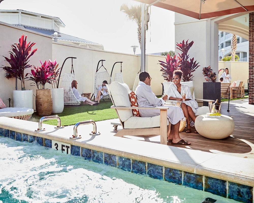A sunny poolside patio with lounge chairs, potted plants, and people relaxing and chatting under shade, enjoying a resort vibe.
