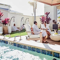 A sunny poolside patio with lounge chairs, potted plants, and people relaxing and chatting under shade, enjoying a resort vibe.