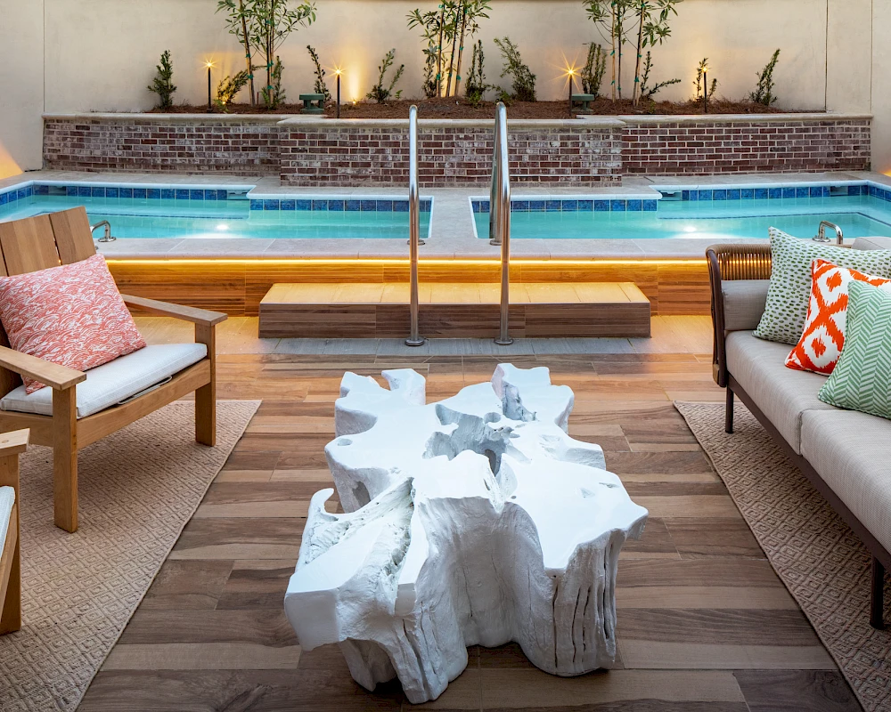 A stylish poolside lounge with wooden deck, cushioned seating, coral pillows, and a unique white rock-like coffee table near a swimming pool.