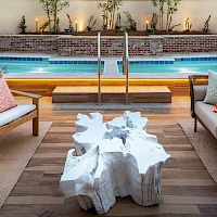 A stylish poolside lounge with wooden deck, cushioned seating, coral pillows, and a unique white rock-like coffee table near a swimming pool.