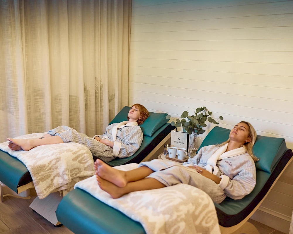 Two people relax on spa loungers, heads back, feet up, with blankets, in a serene, softly lit room.