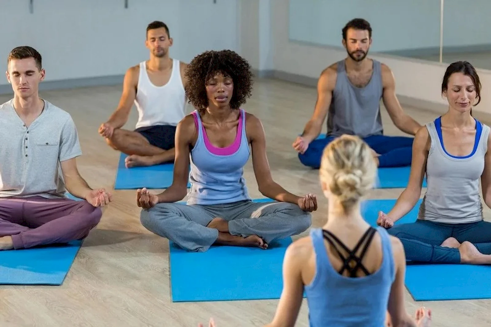 A diverse group sits cross-legged on blue mats in a yoga class, eyes closed, meditating as the instructor leads from the front.