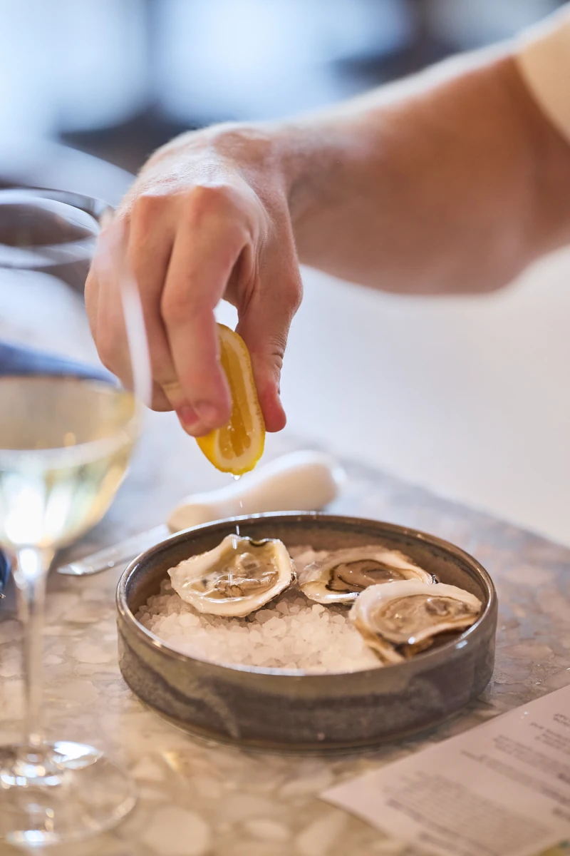 A hand sprinkles lemon on a plate of oysters on ice, with a glass of white wine nearby, in a casual dining setting.