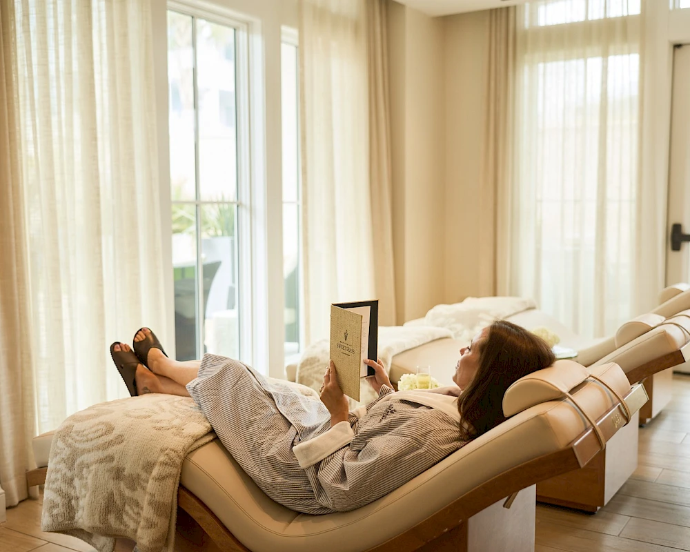 A person lounging on a reclining chaise, reading a tablet beside a bright windowed room with sheer curtains and soft decor.