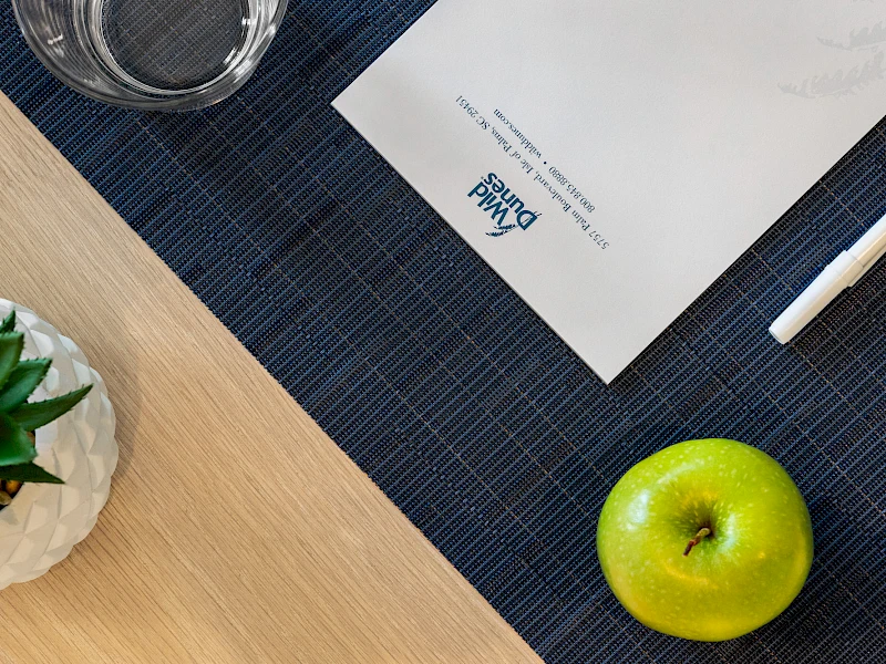 A workspace with a blue placemat, white envelope, pen, a green apple, a small plant, and a glass on the side, all arranged neatly.