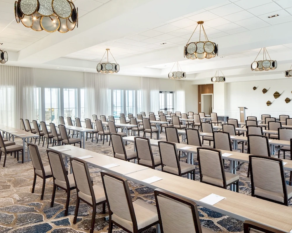 A large conference room with rows of desks and chairs, set for a meeting or seminar, bright windows, and decorative ceiling lights.