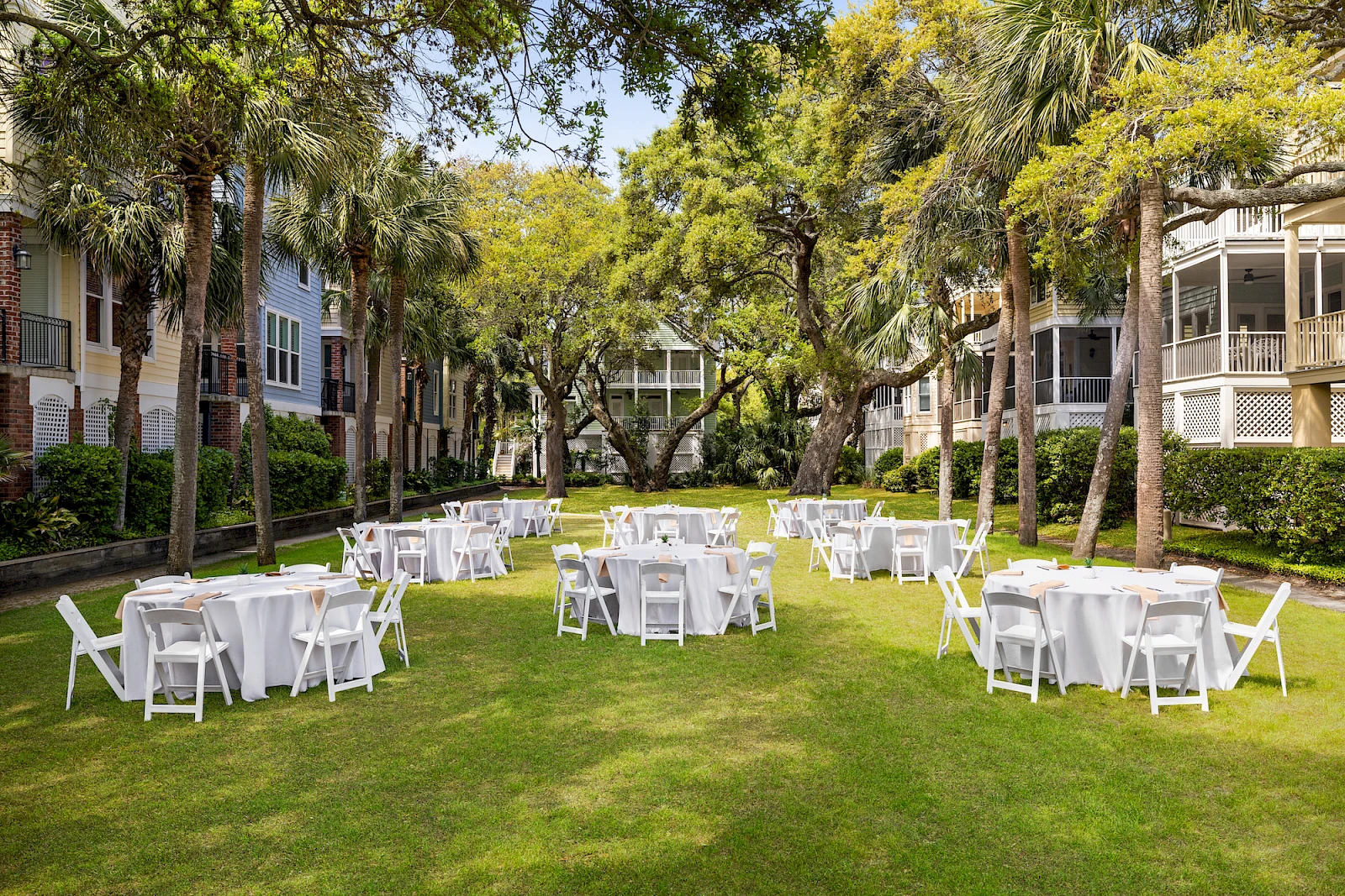 An outdoor lawn setup with white round tables and chairs arranged under trees, likely prepared for an event, on a sunny day.