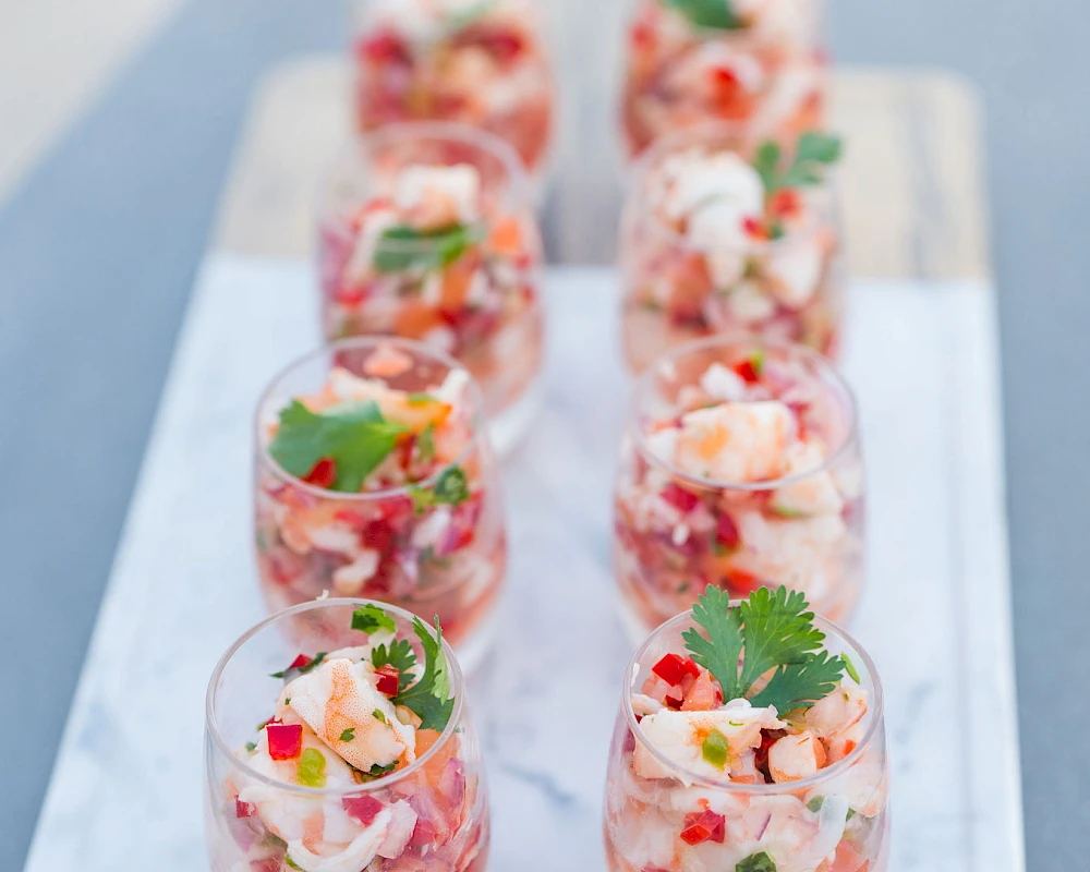 A tray of colorful shrimp cocktails or shrimp skewers arranged in small glasses, garnished with herbs and diced vegetables, ready to serve.