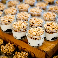 A countertop display of layered dessert cups with cream, cookies, and crunchy granola topping in small glass jars, arranged neatly.