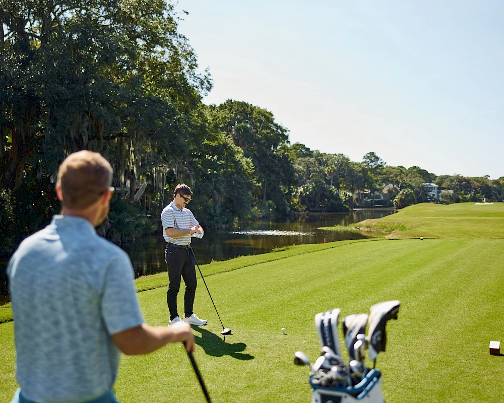 Two golfers on a sunny course, one in the foreground swinging, with a bag of clubs nearby and a calm lake in the background, a peaceful scene.