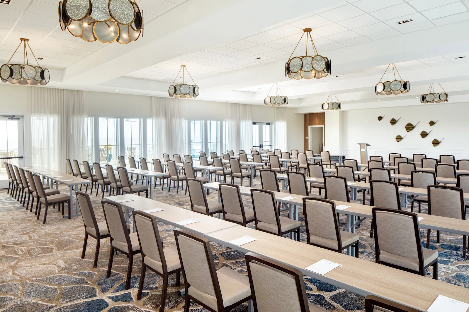 A large conference room with long rows of beige chairs and light wood tables arranged theater-style, modern chandeliers, and windows along one side, empty and ready for a meeting. Top it at 140 characters, always ending the sentence.