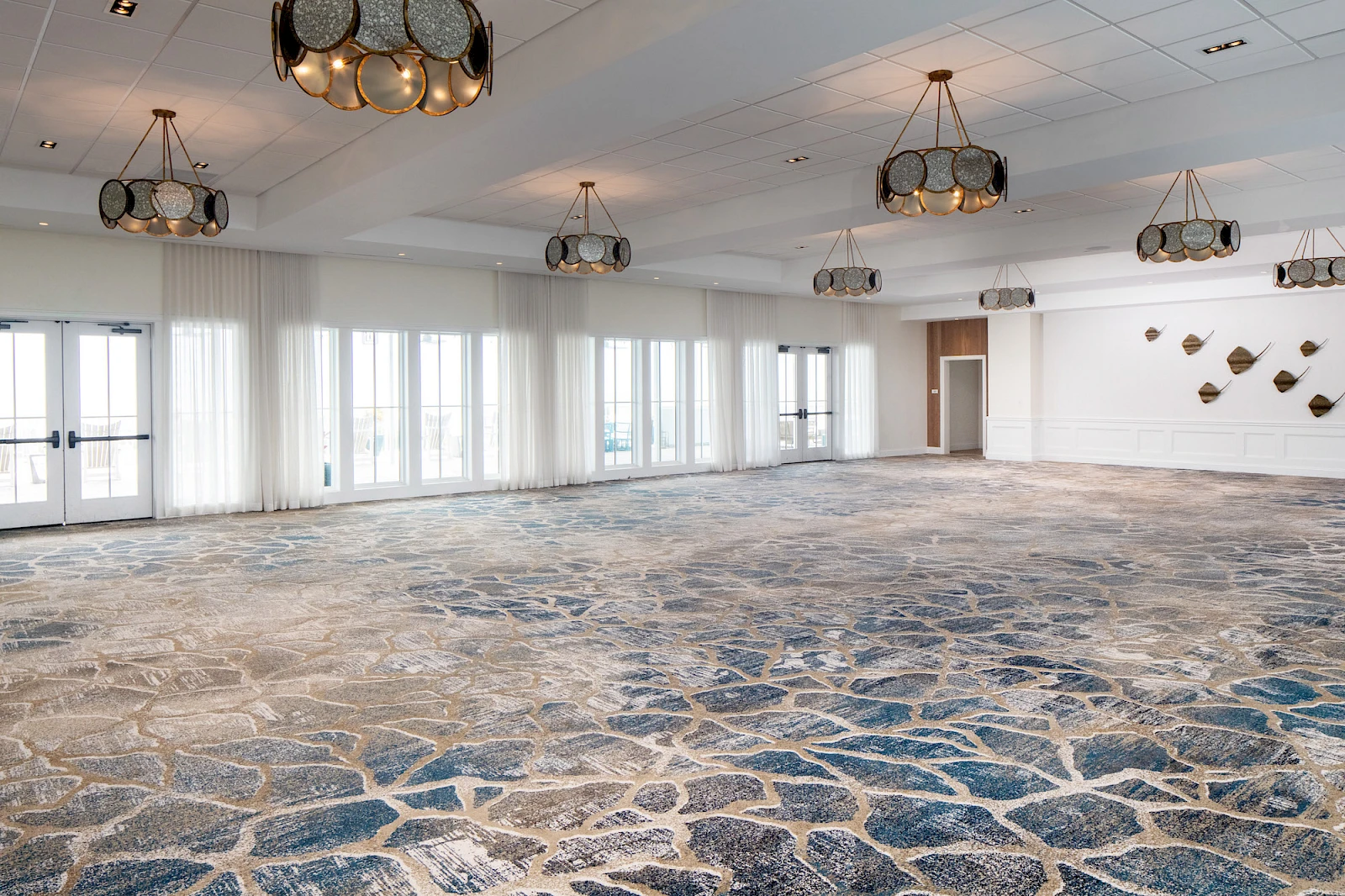 A large, bright empty event room with patterned stone-like carpet, multiple pendant lights, and windows along the left wall.
