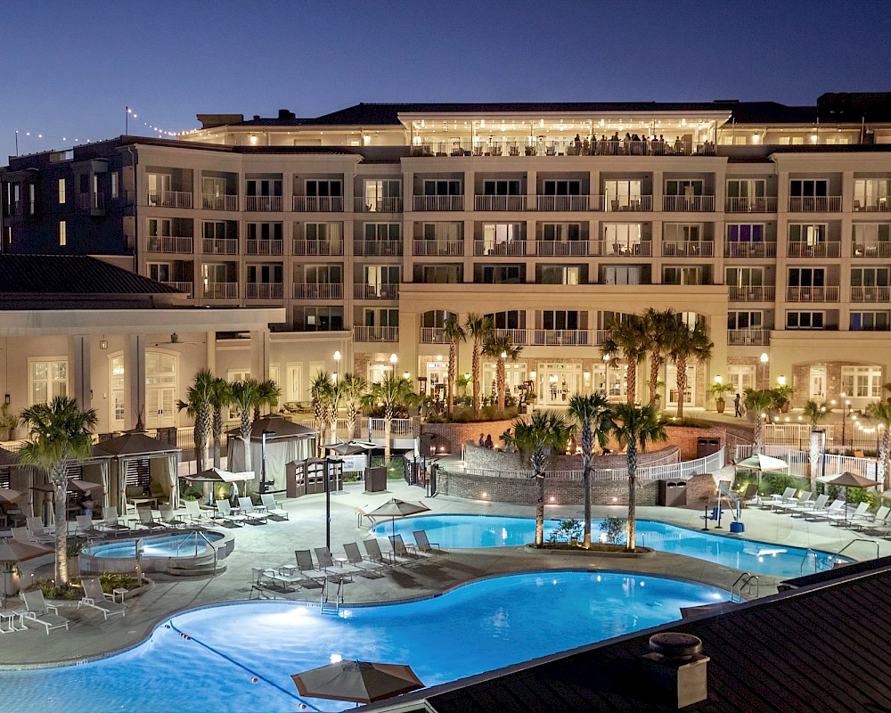 A luxury hotel poolside at dusk, with illuminated infinity-style pool, palm trees, lounge chairs, and a grand multi-story building in the background.