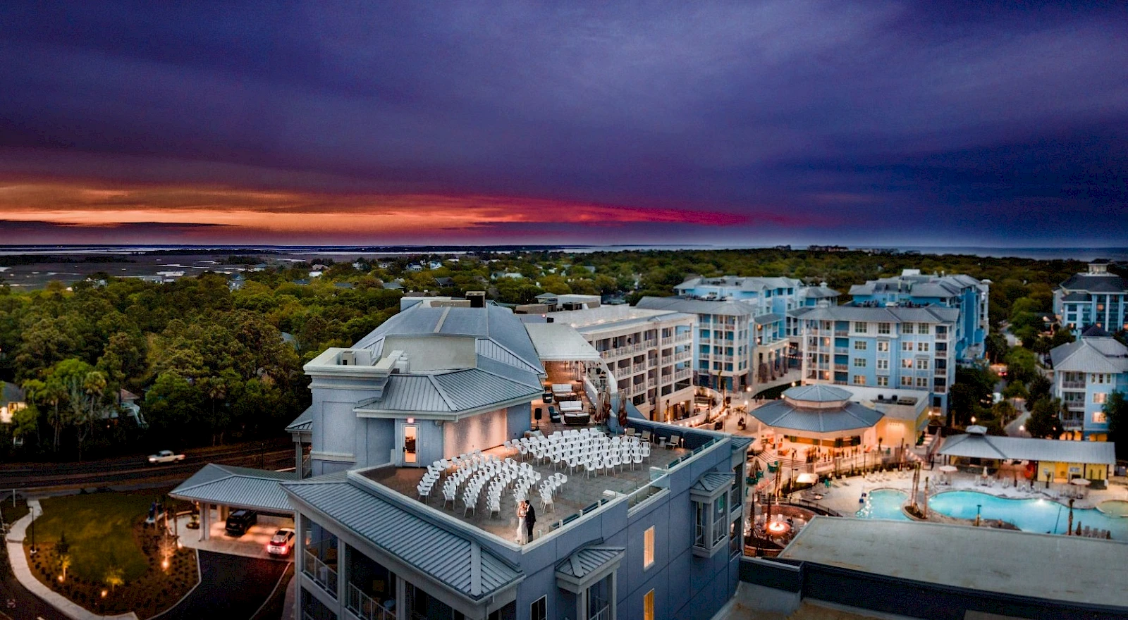 Aerial view of a resort complex with modern buildings, a central pool area, and a sunset over a tree-lined backdrop, tranquil and scenic.